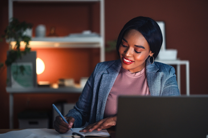 A confident woman in a modern home office space, surrounded by shelves and warm lighting, writing notes energetically while sitting at her desk, projecting professionalism and productivity.